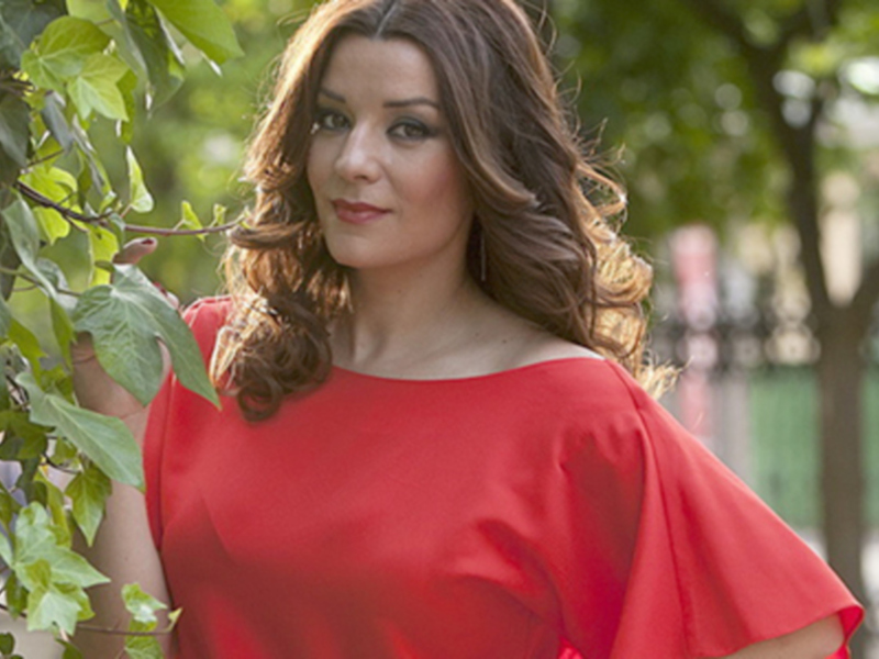 Una joven mujer con cabello largo y ondulado, vestida con un elegante vestido rojo, posa frente a una rama de árbol verde. La escena transmite un aire romántico y relajado, ideal para una tarde soleada en el parque.