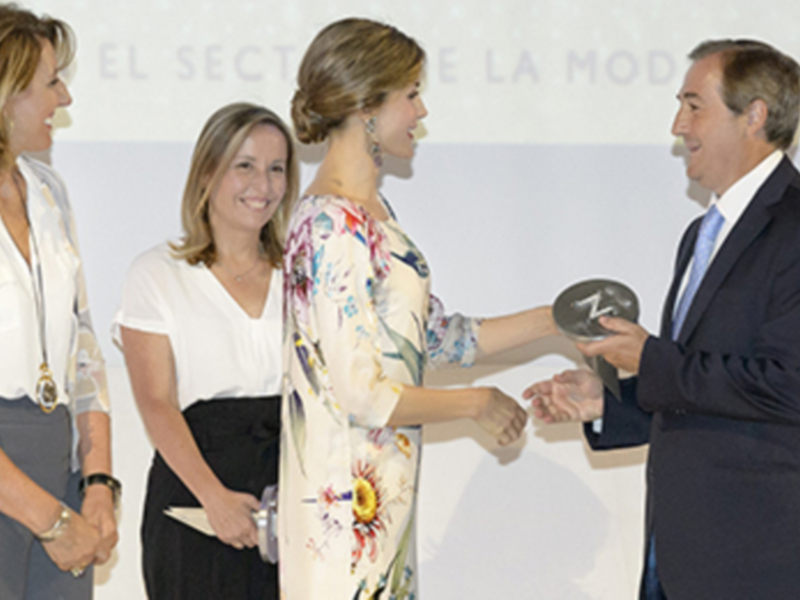 Una mujer recibe un premio de manos de un hombre, mientras una mujer observa la escena.