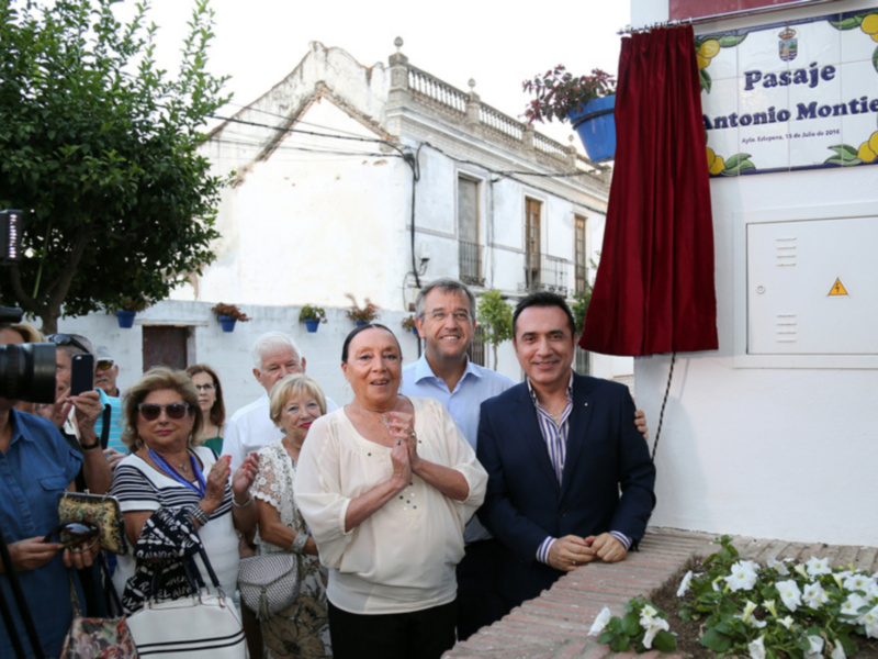 Una familia feliz posa frente a una casa blanca con un letrero que dice "Pasaje Antonio Montiel". La imagen muestra a la familia sonriendo y posando para una foto, con un hombre en el centro. En el fondo se puede ver una casa blanca con detalles de madera y un letrero rojo. La escena parece ser en una calle o plaza, con plantas y un árbol a la izquierda.