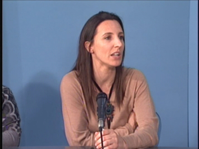 Mujer hablando en un evento con micrófono en mano, fondo azul claro.