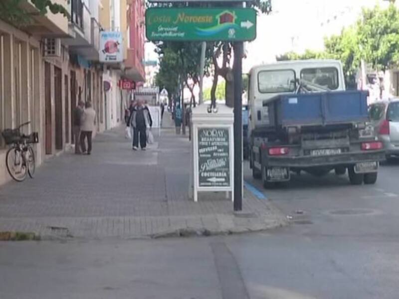 Se observa una calle con edificios a ambos lados, un letrero verde indicando dirección hacia "Noreste", y una bicicleta apoyada en un poste. También se pueden ver vehículos estacionados y personas caminando por la calle.