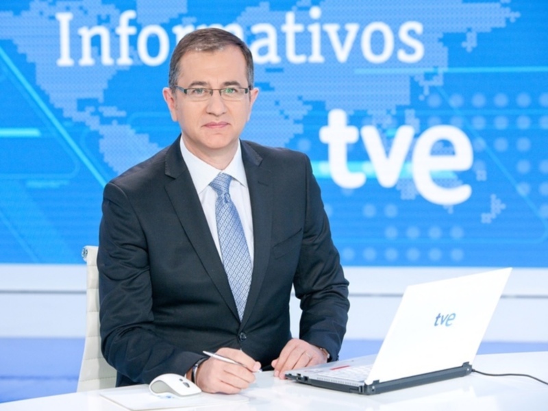 Un presentador de noticias en un estudio de televisión, con una computadora y un marcador sobre la mesa. El fondo muestra el logo de TVE y la palabra 'Informátivos'.