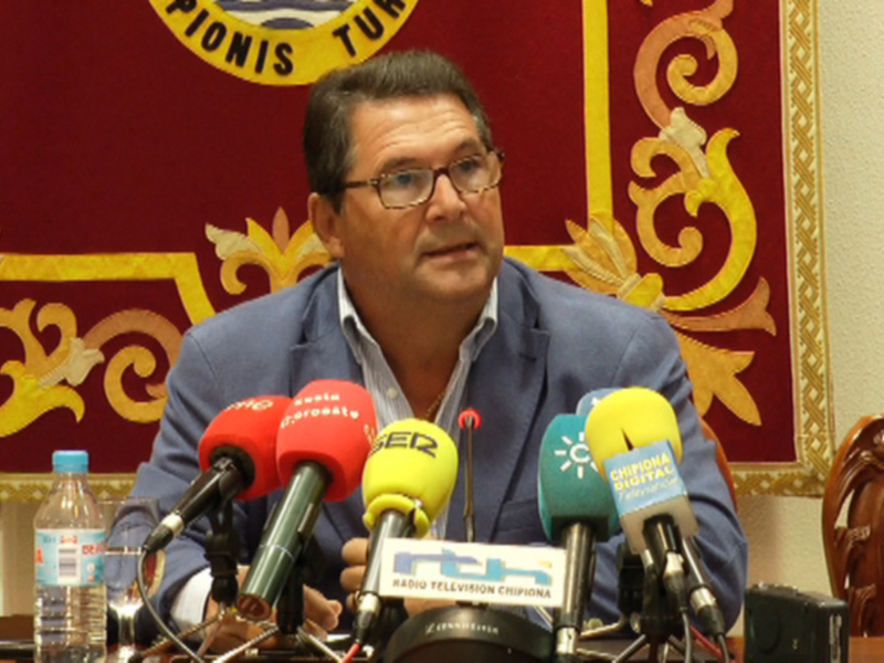 El hombre está en una conferencia de prensa, hablando frente a varios micrófonos de diferentes emisoras. La pared detrás tiene un escudo con símbolos y texto en el centro, probablemente representando una institución o organización.