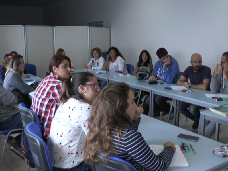 Alumnos en una clase, con libros y lápices en la mesa. La sala tiene una pizarra en el fondo.