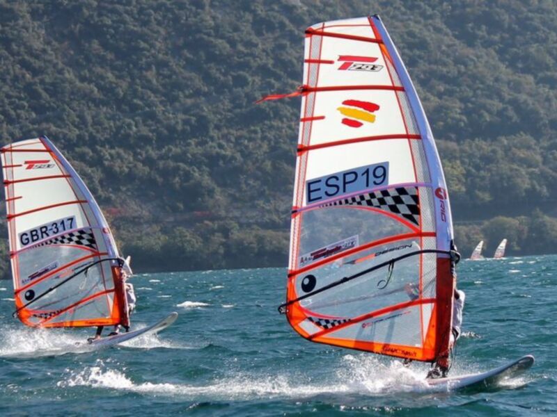Vela de windsurf con la bandera de España en el agua azul con montañas verdes en el fondo.