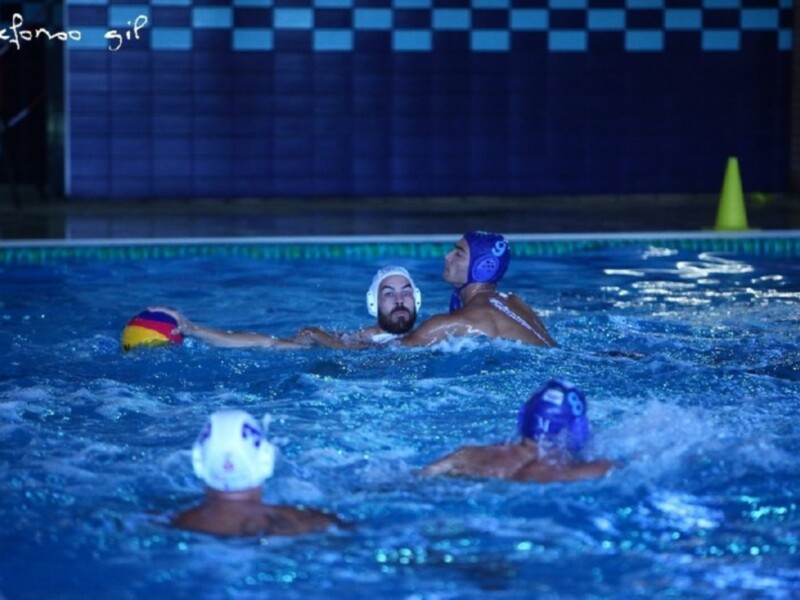 Jugadores de waterpolo en acción en una piscina interior, con cascos azules y un balón rojo y amarillo. El agua es azul y brillante, y hay una línea de división clara en el fondo de la piscina.