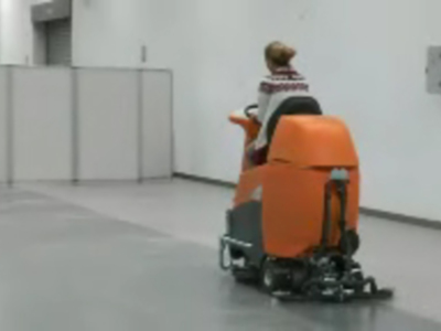 Una persona en una máquina de limpieza de suelo en un entorno industrial. La máquina es grande, con un color naranja y gris, y tiene ruedas para moverse. La persona está sentada en la máquina, mirando hacia el frente. El entorno es limpio y se parece a un almacén o fábrica.