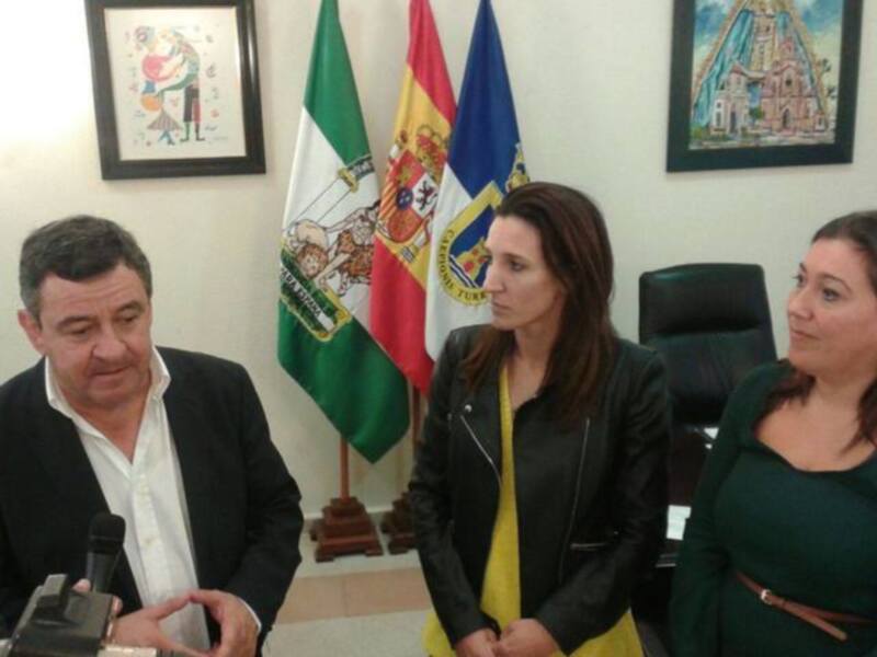Se observa a tres personas en un entorno formal, posiblemente una conferencia o reunión. En el fondo, se pueden ver banderas y cuadros decorativos, lo que sugiere un ambiente institucional. La persona en el centro parece estar haciendo una declaración o presentación, mientras que las dos personas a sus lados escuchan atentamente.
