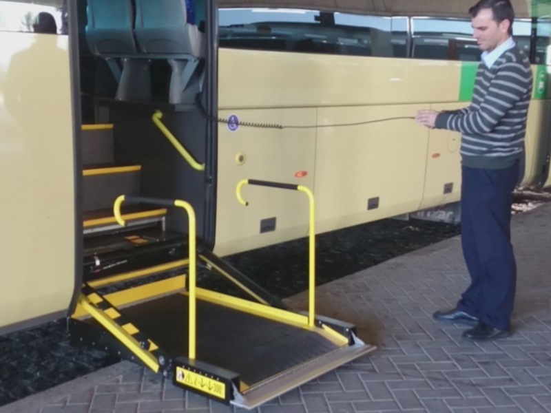 Un hombre está usando una rampa de acceso para discapacitados en un autobús amarillo. La rampa está abierta y el hombre parece estar preparando para subir o bajar del autobús. El autobús está estacionado en una zona de aparcamiento, y hay otros vehículos al fondo.