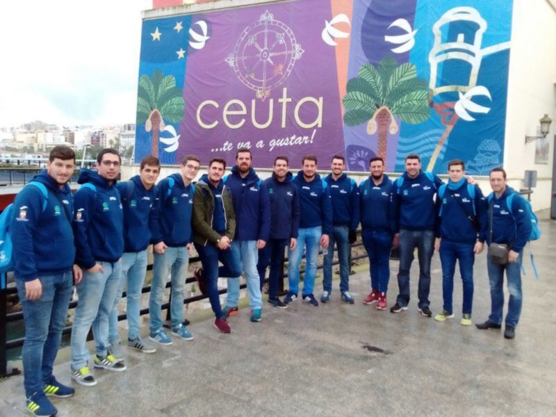 Un grupo de jóvenes en uniforme azul se encuentra frente a un mural con el texto "ceuta" y una bandera de Ceuta. El mural incluye elementos como palmeras, un globo y símbolos de Ceuta.