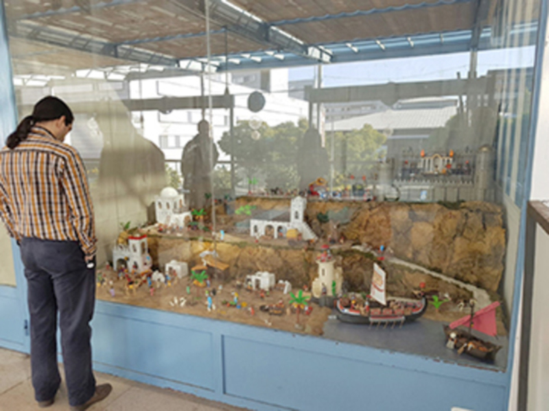 Una persona observa una maqueta de un pueblo en un vitrín. La escena muestra casas, animales y barcos de juguete en un paisaje montañoso. La vista exterior muestra árboles y edificios en la distancia.