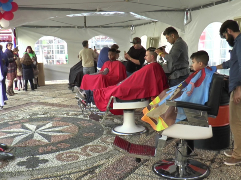 Evento de barbería al aire libre con clientes y peluqueros en una carpa decorada con globos.