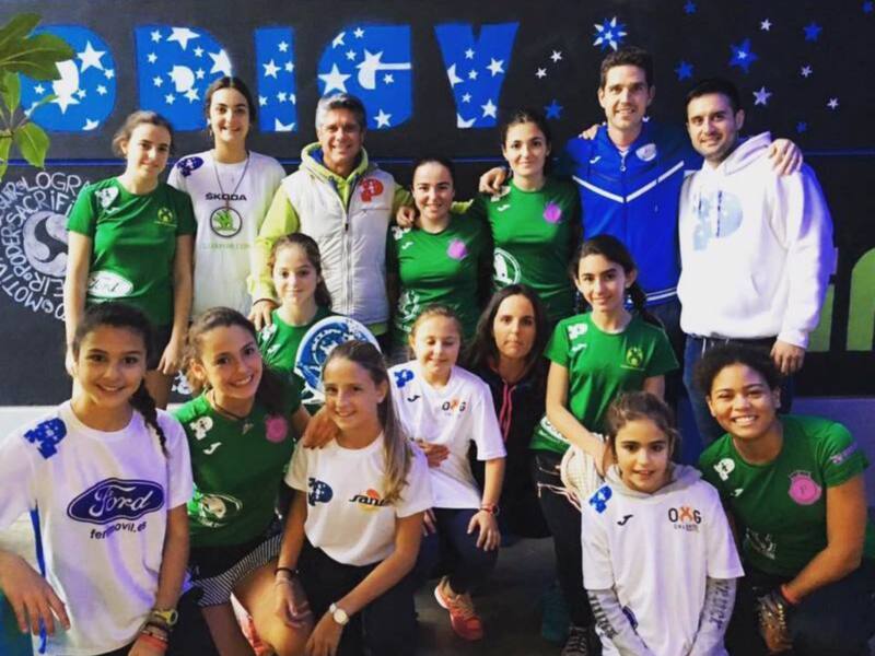 Equipo de fútbol femenino posando para una foto en el estadio. Las jugadoras lucen camisetas de diferentes equipos, y el fondo muestra el nombre del club deportivo. Los entrenadores y algunos familiares también están presentes en la imagen.