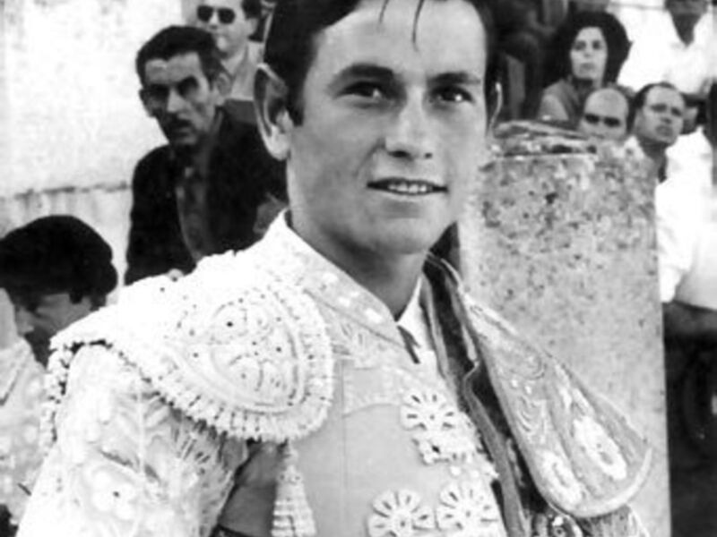 Imagen en blanco y negro de un torero en traje de cuero, con una capa de terciopelo y un sombrero de copa. El fondo muestra a personas en el público, algunas con sombrillas. El torero tiene una expresión seria y está mirando hacia la cámara.