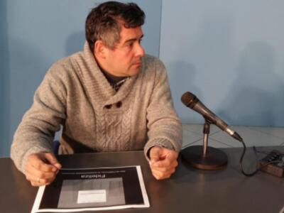 Un hombre se sienta frente a un micrófono en una sala de conferencias, con un documento en la mano izquierda. El fondo es azul y la iluminación es suave.