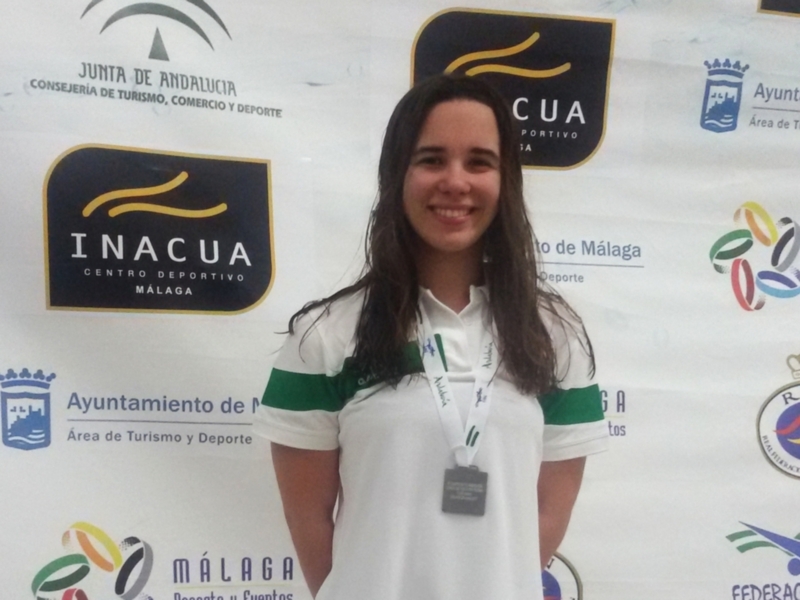 A una persona sonriendo frente a un fondo con logos de la Junta de Andalucía, Consejería de Turismo, Comercio y Deporte, CUA (Consejería de Cultura y Juventud), Ayuntamiento de Málaga, y la Federación Española de Deportes Acuáticos. La persona lleva una camiseta blanca con detalles verdes y un cuello verde, junto a una medalla.