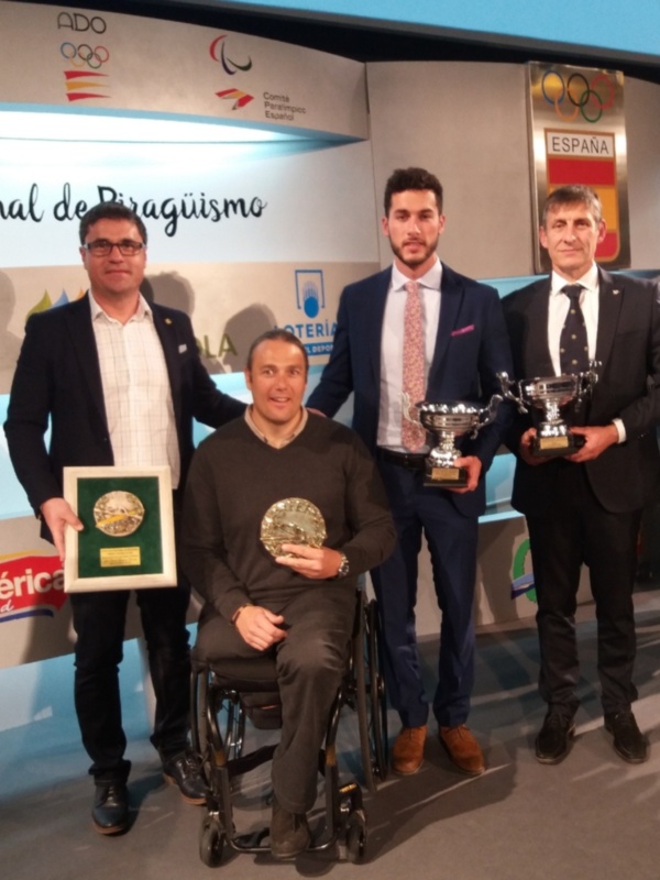 Tres hombres, uno en silla de ruedas, posan con trofeos y medallas en un evento deportivo.