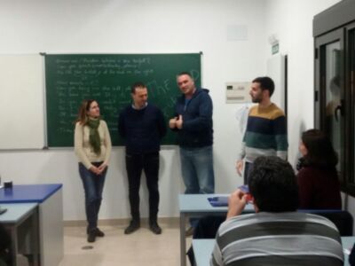 En una aula de clase, un grupo de personas se encuentra en frente de una pizarra con escritos matemáticos. La escena muestra a dos hombres y una mujer en el centro, con otros estudiantes sentados en las mesas al fondo. La pizarra es la principal fuente de luz, iluminando a todos los presentes.