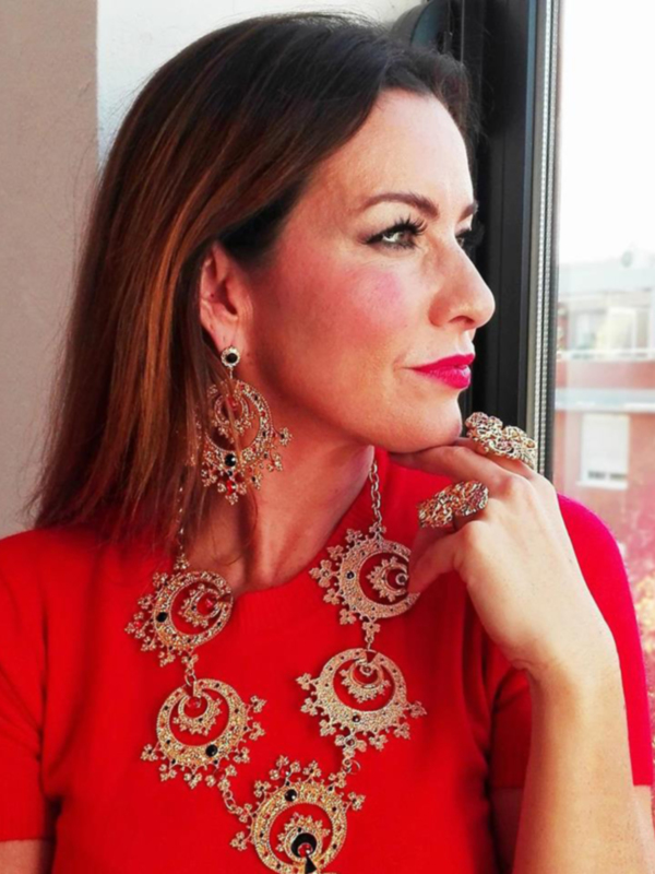 Una mujer con cabello largo y rojo, vestida con un vestido rojo con bordados dorados, posa con una mano en la barbilla, frente a una ventana con una vista de edificios en la distancia.