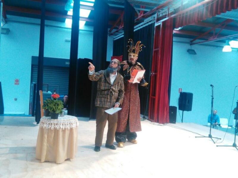 Dos personas en trajes de época, uno con sombrero y el otro con traje largo, presentan un documento en un escenario con iluminación azul y roja.