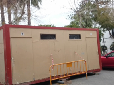 Un pequeño edificio móvil de color beige con una cubierta roja está estacionado en la calle, rodeado por árboles y una valla amarilla.