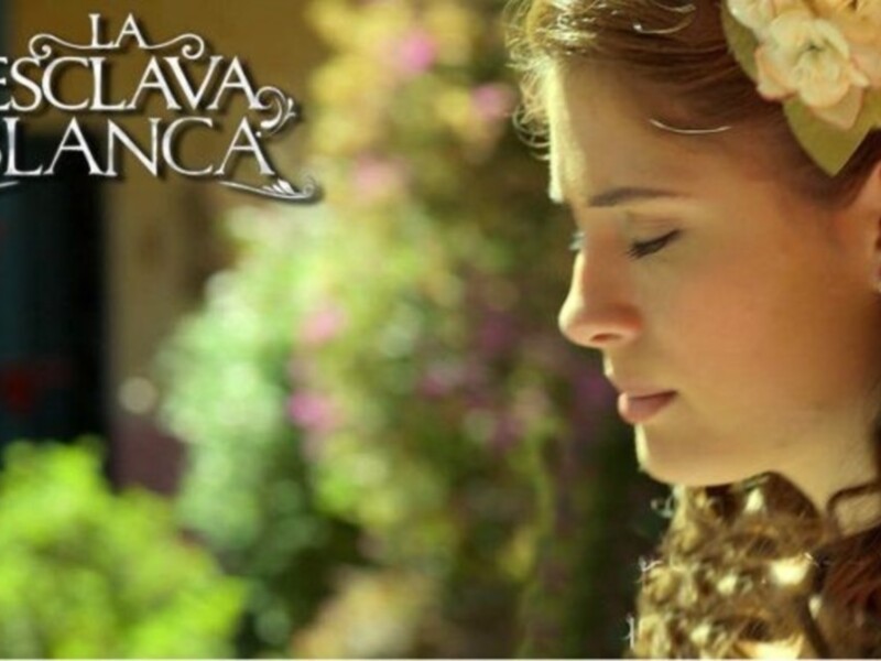 A una joven con cabello largo y rizado, adornada con un diadema de flores amarillas y rosadas. La escena parece ser al aire libre, con un fondo borroso de vegetación y flores.