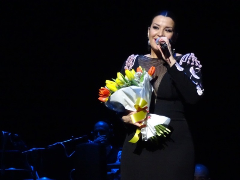 Una mujer en un concierto sostiene una hermosa guirnalda de flores, incluyendo amarillas y rojas. Está vestida con un elegante abrigo negro con detalles en blanco, posiblemente de seda. La iluminación es dramática, enfocándose en ella y el ramo de flores.