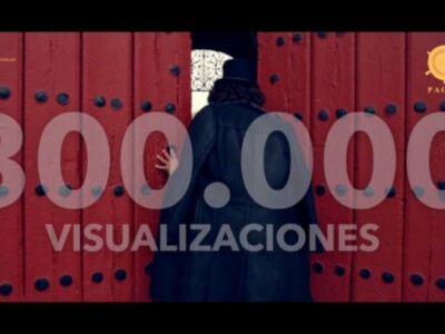 Una persona con un sombrero negro y una capa negra se acerca a una puerta roja con numerosas bisagras. La imagen muestra el número 300000000 en letras blancas, y debajo se lee "VISUALIZACIONES". La imagen es parte de un anuncio o comercial.