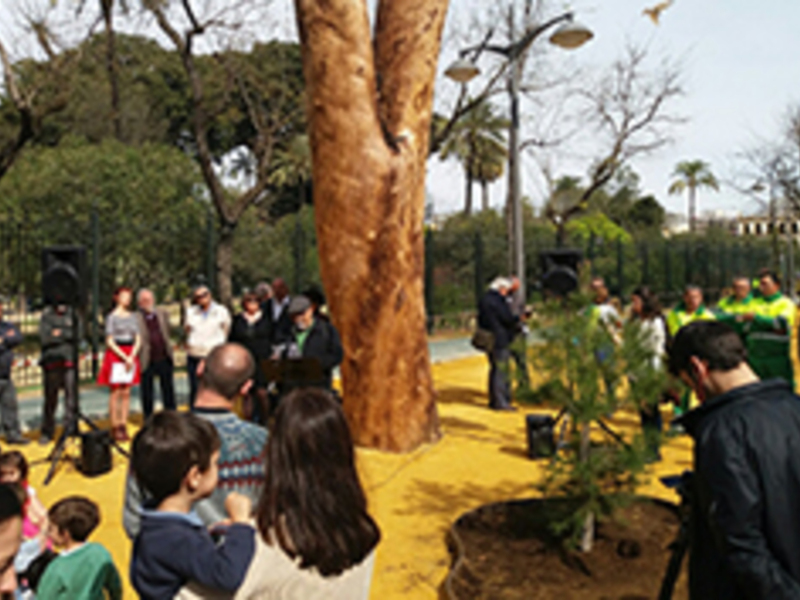 Una multitud de personas, incluyendo niños y adultos, observan con atención una actividad en un área verde. Algunos están tomando fotos o mirando hacia arriba, mientras otros conversan entre sí. La escena se desarrolla en un entorno urbano con árboles y una estructura de madera visible. La atmósfera es tranquila, con un cielo despejado que sugiere una jornada soleada.