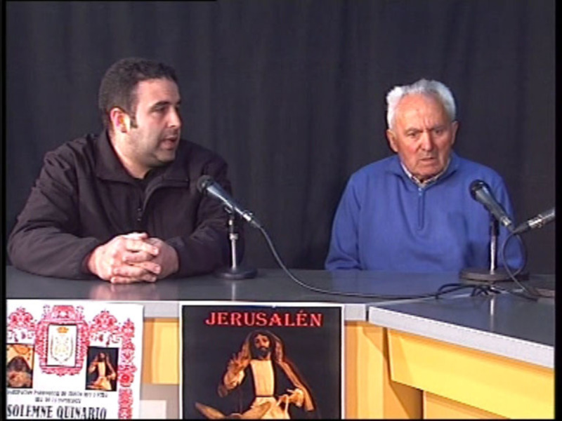 Dos hombres sentados en una mesa con micros, uno de ellos hablando mientras el otro escucha. En el fondo, hay una pantalla con imágenes religiosas y el texto 'Jerusalén'.