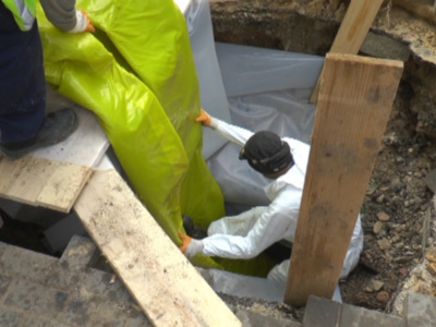 Un trabajador en traje de protección y guantes se desliza por una escalera de madera para acceder a un hueco en el suelo. La escena muestra una construcción o reparación de edificios antiguos, con bloques de piedra y estructuras de madera.