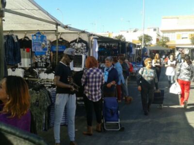 En una calle peatonal, varias personas caminan y conversan mientras pasan por un mercado de ropa. El cielo es azul claro, y a lo lejos se pueden ver edificios y árboles. La escena es relajada, con gente de diferentes edades y estilos vestidos.
