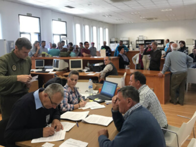 Una sala de reuniones con varias personas sentadas en mesas, trabajando y conversando. Hay computadoras, escritorios y libros en la mesa central. La pared está llena de ventanales que permiten la entrada de luz natural. El ambiente es profesional y ordenado.