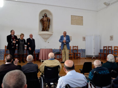 Una sala de conferencias con una estatua en el centro y un cartel en la pared. Personas sentadas en sillas, algunos en sillas de ruedas, escuchando a tres personas que están hablando.