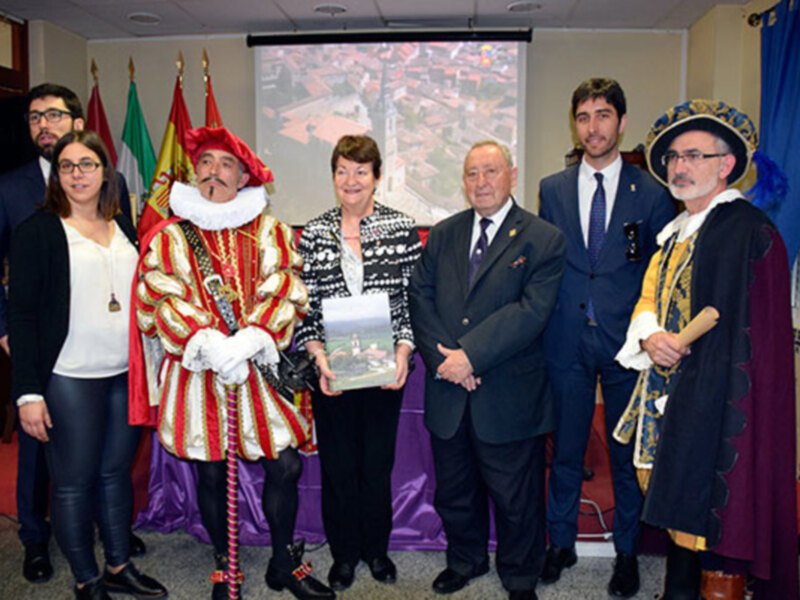 Una reunión formal con personas vestidas de manera distintiva, incluyendo un personaje histórico en traje rojo y dorado, posando para una foto en un salón con banderas y una proyección de una ciudad en el fondo.