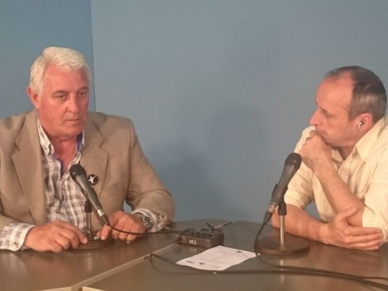 Dos hombres sentados en una mesa con micrófonos, uno de ellos parece estar en una entrevista o conferencia de prensa.