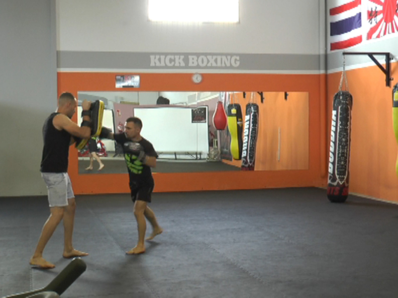 Un hombre realiza un entrenamiento de boxeo en una sala de fitness con paredes naranjas y grises. En la pared, hay un cartel que dice "KICK BOXING" y una bandera de Tailandia. El suelo es de color gris oscuro, y hay un saco de boxeo colgando en la pared.