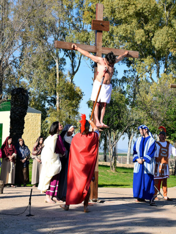 Una escena de la Pasión de Cristo, con una persona en la cruz, rodeada de personas vestidas con trajes históricos, en un entorno natural con árboles y un cielo despejado.