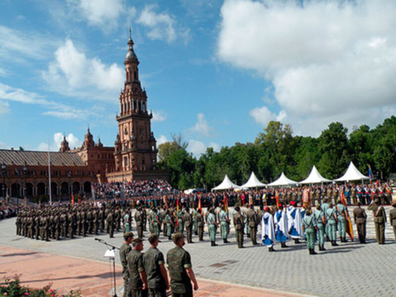 Una imagen de un desfile militar en una plaza con militares uniformados y banderas ondeando. En el fondo, se puede ver la Torre de la Plata en Sevilla, España. El cielo está despejado con algunas nubes.