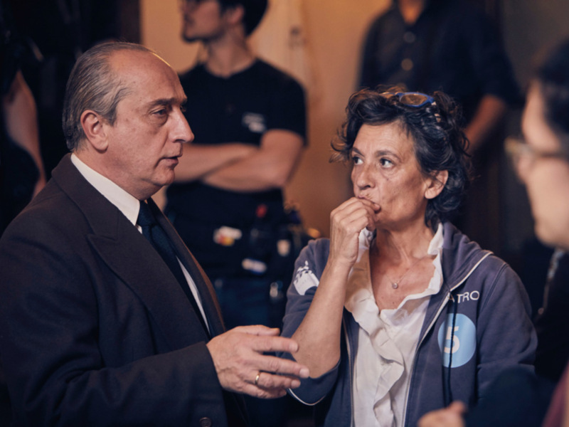 Dos personas conversan en un entorno formal, uno de ellos con una camiseta azul y otro con traje. La escena sugiere un evento o reunión importante, posiblemente relacionado con la industria de los medios o el entretenimiento.