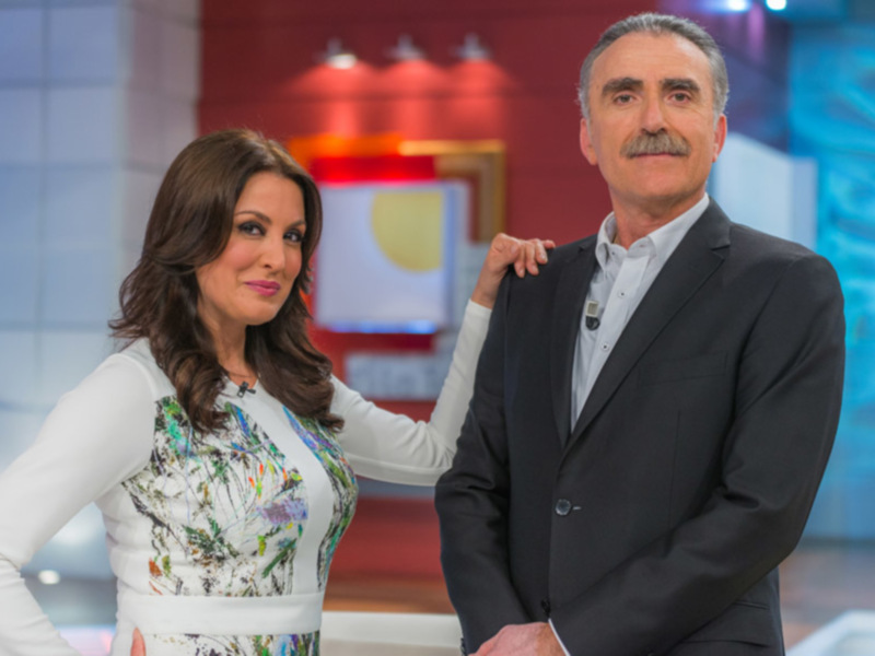 Dos personas sonrientes en un estudio de televisión. La mujer lleva una camisa blanca con estampado floral y el hombre está vestido con un traje negro. El fondo es de color rojo y azul, con luces en el techo.