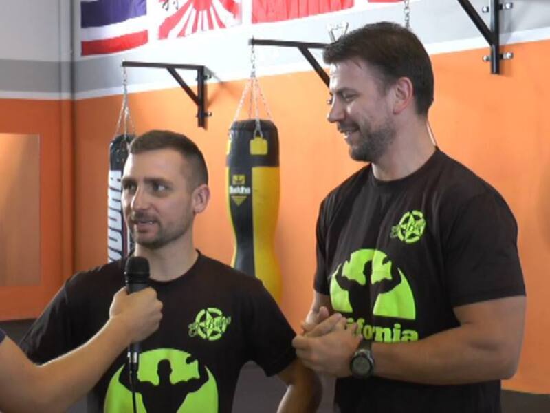 Entrevista en un gimnasio con dos hombres, uno con micrófono y otro con una camiseta negra con logotipo verde.