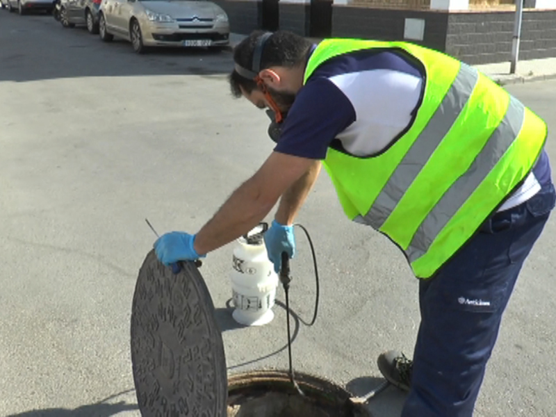 Un trabajador en un chaleco reflectante de color verde está limpiando una alcantarilla con una aspiradora. El fondo muestra un camino asfaltado y vehículos estacionados en el lado opuesto.
