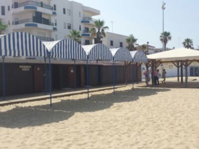 Barras de playa con toldos azules y blancos, frente a edificios blancos. Poca gente en la arena.