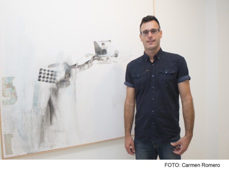 Un hombre posa junto a una obra de arte que parece ser un retrato abstracto, con detalles que podrían representar un mapa y una bandera. La imagen está firmada por Carmen Romero.