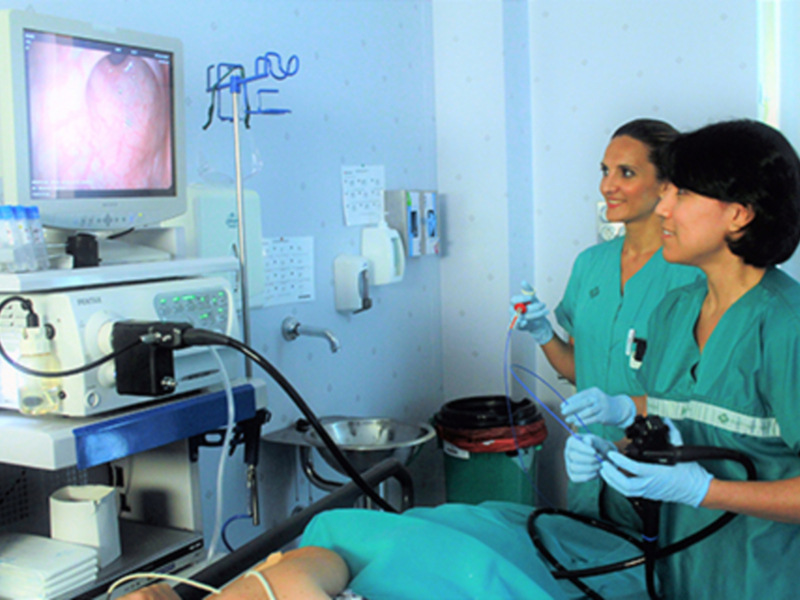 Dos médicos en una sala de operaciones, uno con un monitor que muestra una imagen interna del cuerpo humano. La escena está en un hospital, con equipos médicos y una silla de ruedas.