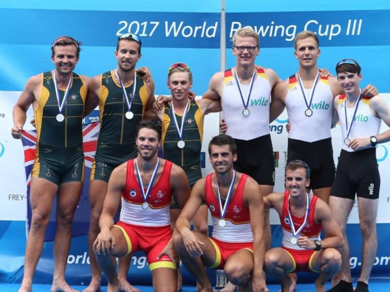 Equipo de remo celebrando su victoria en el 2017 World Rowing Cup III, con medallas en el pecho y una bandera de la Unión Europea en el fondo.