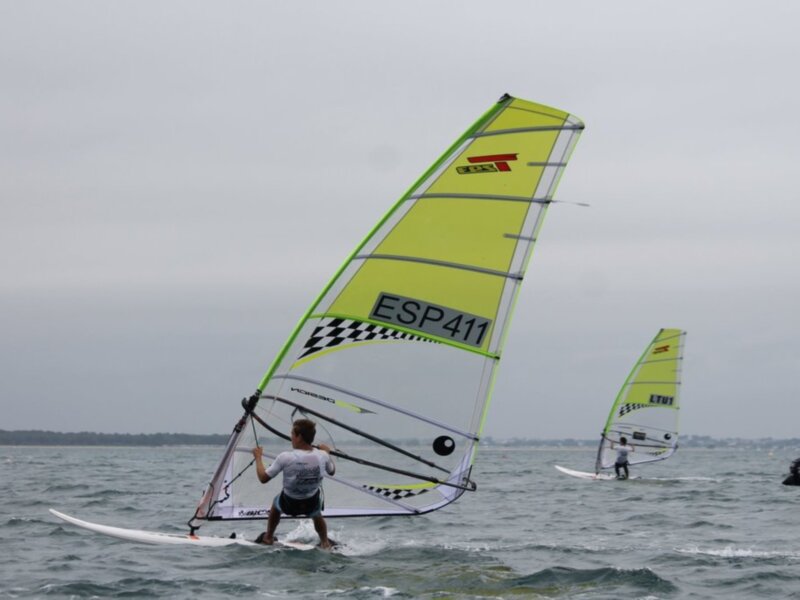 Two windsurfers on a cloudy day, with one in a bright yellow sail and the other in a white sail with a black and white checkered pattern.