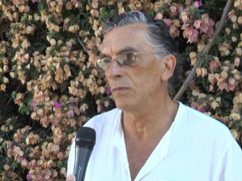 Un hombre con gafas y una camisa blanca está hablando en un micrófono frente a una planta con flores coloridas.