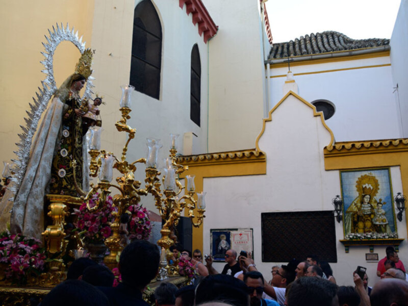 Procesión religiosa con imagen de la Virgen en un entorno de iglesia con decoración floral y candelabros dorados.